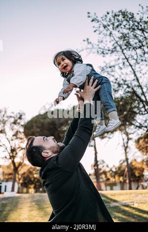 Vater hat Spaß mit seiner Tochter im Park, die sie in die Luft wirft, während sie glücklich lacht. Stockfoto