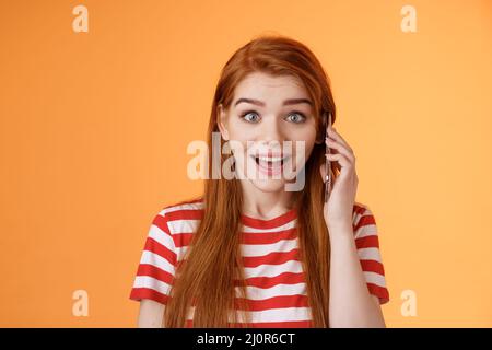 Freuen Sie sich auf die Stimme von Freunden, die im Ausland anrufen. Amüsiert überrascht niedlich Rotschopf fröhliche Mädchen abholen Telefon, halten Smartphone-Ohr, finden ou Stockfoto