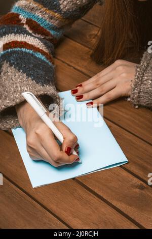 Frauenhand mit roter Maniküre auf einem Holztisch Schreiben Sie eine Grußkarte mit einem Stift. Frau füllt einen Brief auf einem Stück blauer Pape aus Stockfoto