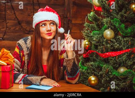 Charmante Frau in einem roten Hut mit weißem Fell sitzt an einem Tisch in der Nähe eines geschmückten Weihnachtsbaums und denkt, was man dem Weihnachtsmann schreiben soll. Cl Stockfoto