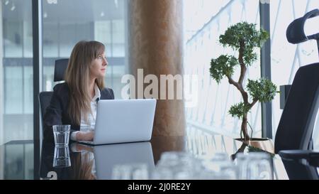 Glückliche junge erfolgreiche weiße Geschäftsfrau, die im Büro am Schreibtisch sitzt, denkt, mit einem Laptop beschäftigt ist und lächelt. Stockfoto