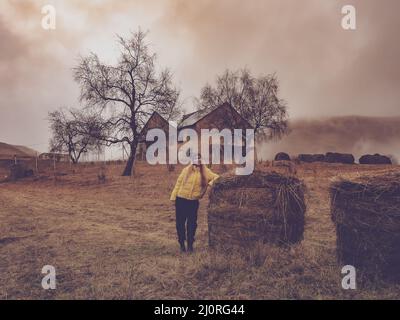 Ein Mädchen steht in einer verlassenen Landschaft zwischen Heuhaufen vor dem Hintergrund eines verlassenen alten Hauses in einem mystischen Dunst aus Wolken Stockfoto