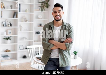 Hübscher Junger Arabischer Kerl, Der Zu Hause Mit Gefalteten Armen In Der Nähe Des Schreibtisches Steht Stockfoto