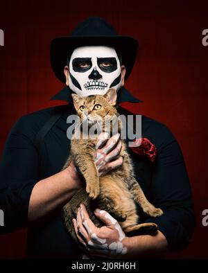 Ein Mann in Skelett-Make-up hält eine Erwachsene Katze auf einem roten Hintergrund in seinen Armen. Halloween-Party Stockfoto