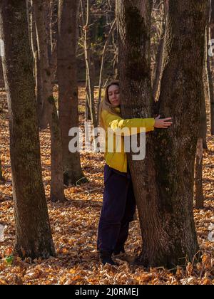 Eine Frau steht an einem sonnigen Herbsttag in einem dichten Wald inmitten von abgefallenen Blättern und umarmt moosigen Baumstamm Stockfoto