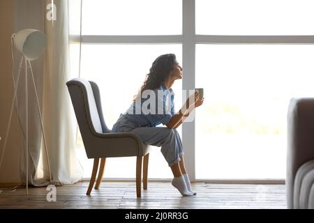 Seitenansicht einer schönen jungen Frau, die heißen Kaffee trinkt, auf einem Stuhl neben dem Fenster zu Hause sitzt, freier Platz Stockfoto