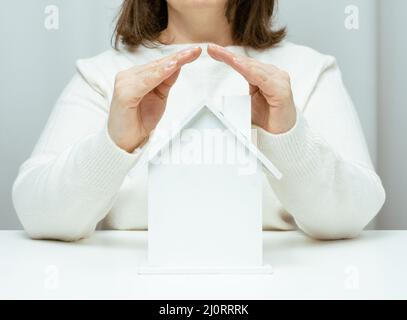 Weibliche Hände gefaltet zueinander über einem hölzernen Miniatur-Modellhaus auf weißem Hintergrund. Immobilienversicherungskonzept, envi Stockfoto