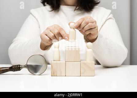 Eine Frau in einem weißen Pullover legt eine Holzfigur auf das Podium. Das Konzept der Mitarbeitersuche, Karriereentwicklung. Talentierte em Stockfoto