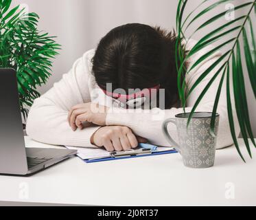 Frau in einem Pullover schläft an einem Arbeitstisch neben einem Laptop. Ermüdung und Überarbeitungs-Konzept. Faulheit Stockfoto