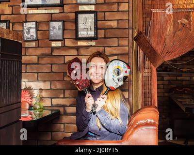 Russland, Kislowodsk 05.11.2021. Lächelnde Frau hält halloween-Masken, während sie im Retro-Café auf dem Sofa sitzt Stockfoto