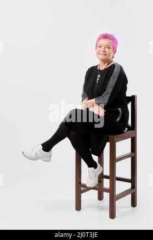 Die alte Frau mit kurzen rosafarbenen Haaren lächelt sanft auf einem hohen Stuhl und hält die Hände gekreuzt. Positives Emotionskonzept. Stockfoto