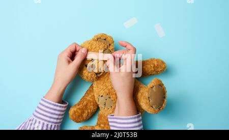 Weibliche Hand hält einen braunen Teddybär und klebt einen medizinischen Klebstoff-Gips auf blauem Hintergrund, Tram-Behandlung Stockfoto