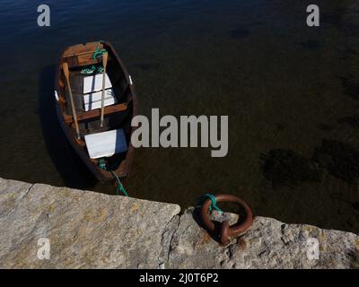 Ein kleines Ruderboot mit den Rudern, die auf dem verwinkelt liegen, vertäut an einem alten Steinkai, einem steinernen Wellenbrecher, in einem kleinen Fischerhafen und Dorf. Stockfoto