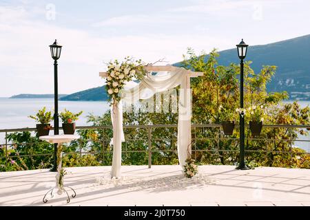 Der mit Rosen und weißen Stoffen verzierte Hochzeitbogen steht am Ufer über dem Meer Stockfoto