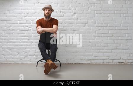 Ernsthafter bärtiger Mann im Hut, der vor einer weißen Backsteinmauer sitzt. Arme gekreuzt. Foto in voller Länge mit Kopierbereich. Stockfoto