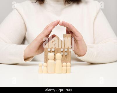 Ein Holzhaus und Figuren einer Familie, zwei weibliche Palmen schützen Objekte von oben. Immobilienversicherungskonzept, glückliche Familie Stockfoto