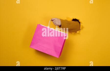 Weibliche Hand mit einer pinken Papiertasche, die aus einem zerrissenen Loch auf einem gelben Papierhintergrund herausragt. Ablehnung aus Kunststoff, Start Stockfoto