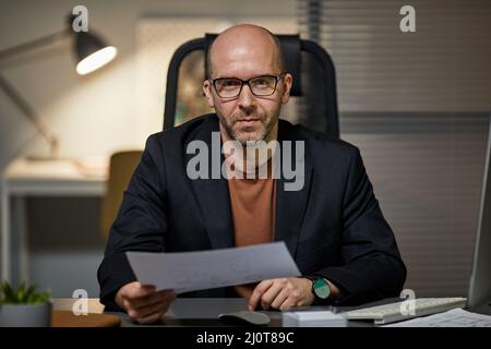 Porträt eines kahlen erwachsenen Geschäftsmannes, der am Arbeitsplatz sitzt und die Kamera anschaut, während er spät im Büro arbeitet Stockfoto