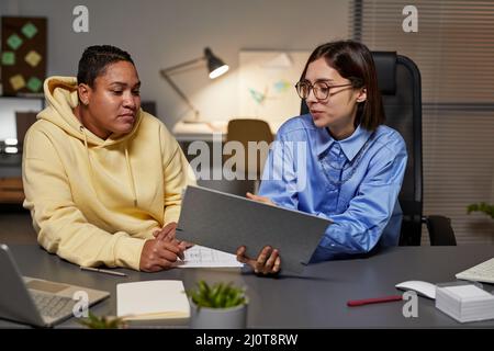 Portrait von zwei jungen Frauen, die während ihrer Arbeit spät in der Nacht im Büro über die Dokumentation diskutieren Stockfoto