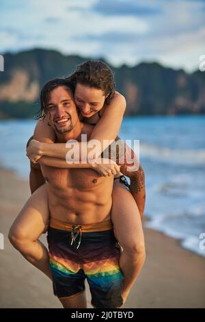 Keine Sorge, sei glücklich. Porträt eines glücklichen jungen Mannes, der seiner Freundin eine Huckepack-Fahrt am Strand gab. Stockfoto