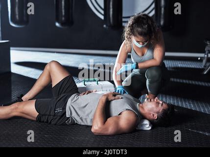 Der Weg zur Gesundheit wurde gerade etwas schwieriger. Schuss einer maskierten jungen Frau, die einem verletzten Mann im Fitnessstudio erste Hilfe geleistet hat. Stockfoto