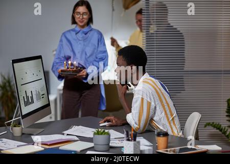 Seitenansicht Porträt eines afroamerikanischen Mannes, der im Büro arbeitet, mit Kollegen, die im Hintergrund Geburtstagskuchen bringen Stockfoto