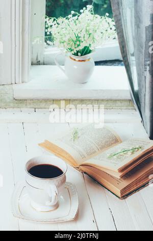 Eine Tasse schwarzen Kaffee und ein offenes Buch auf einem weißen Retro-Holztisch und ein Blumenstrauß von Maiglöckchen auf dem Stockfoto