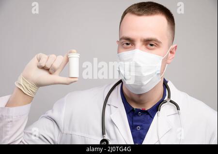 Der junge kaukasische Arzt in einer Maske und einem weißen Mantel hält ein Paket mit unbekannten Pillen in der Hand. Ärztliche Empfehlung von medicati Stockfoto