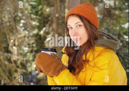 Attraktive kaukasische positive junge Frau in Winterkleidung in einem Hut mit Fäustlingen und einer Daunenjacke steht in einem Nadelgewand Stockfoto