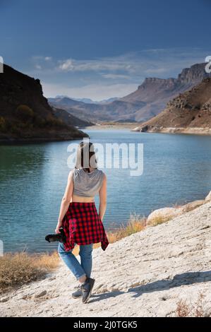 Rückansicht eine Fotografin in legerer Kleidung mit einer professionellen Kamera steht am Ufer eines Sees in den Bergen und l Stockfoto