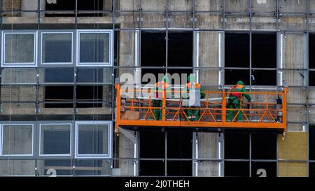 Die Arbeiter in der Bauwiege in der Höhe sind im Winter mit der Verglasung der Fassade eines Wolkenkratzers beschäftigt. Stockfoto