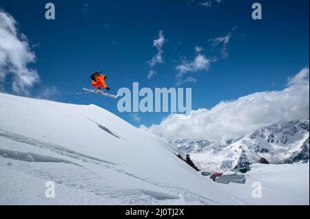 Fliegen in einem Sprung über einen Hang Freestyle Skifahrer springen in einem orangen Skianzug in den verschneiten Bergen an einem sonnigen Tag Stockfoto