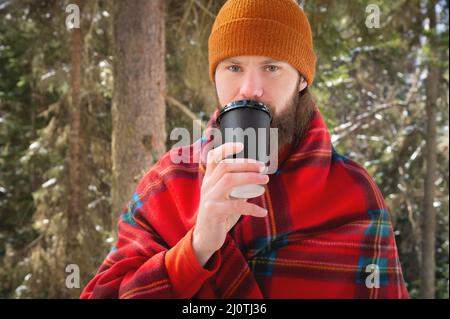 Bärtiger Kaukasusmann in einem Hut, der in eine Decke mit einem Becher heißen Getränks in einem Papierbecher in einem winterlichen Nadelwald auf einem Su eingewickelt ist Stockfoto