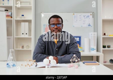 Vorderansicht Porträt eines afroamerikanischen Lehrers, der im Chemieunterricht mit Molekülmodellen am Schreibtisch sitzt, Kopierraum Stockfoto