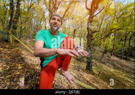 Porträt eines reifen Mannes in einem Schal auf dem Kopf sitzt auf einer straffen Slackline erklärt, wie man im Herbstwald balanciert Stockfoto