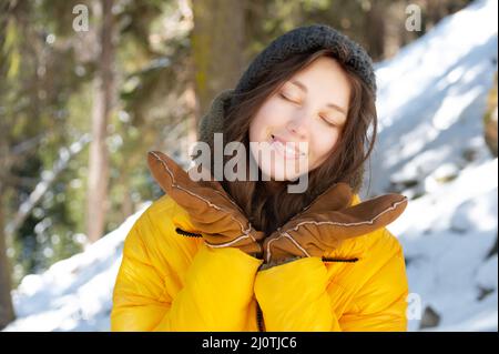 Glückliche junge kaukasische Frau in einer Winter-Daunenjacke Hut und Fäustlinge mit geschlossenen Augen vor Glück freut sich, ihre Hände zu verbreiten Stockfoto