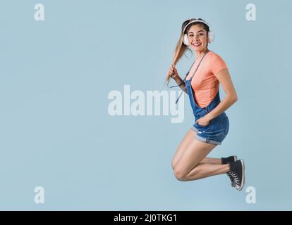 hispanische junge Frau mit Kopfhörern, die springen und Musik auf blauem Hintergrund hören in Mexiko, Lateinamerika Stockfoto