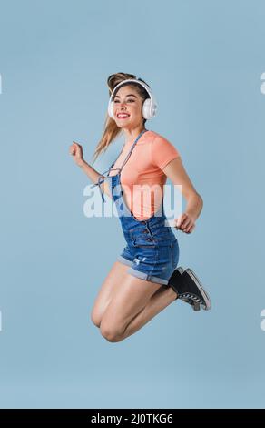 hispanische junge Frau mit Kopfhörern, die springen und Musik auf blauem Hintergrund hören in Mexiko, Lateinamerika Stockfoto