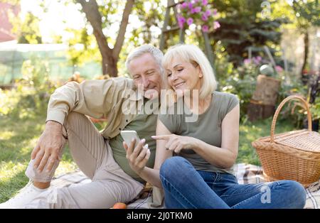 Glückliche ältere Ehegatten sitzen während des Picknicks im Garten auf der Decke und nutzen das Smartphone, um sich am Frühlingstag im Freien auszuruhen Stockfoto