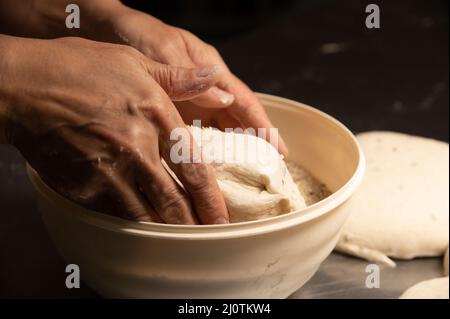 Nahaufnahme der weiblichen Hände einer Bäckerin, die den Teig in eine Schüssel mit Samen und Getreide eintaucht, bevor sie in einer Backstube hausgemachtes Brot backen Stockfoto