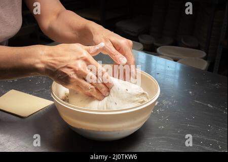 Nahaufnahme der weiblichen Hände einer Bäckerin, die den Teig in eine Schüssel mit Samen und Getreide eintaucht, bevor sie in einer Backstube hausgemachtes Brot backen Stockfoto