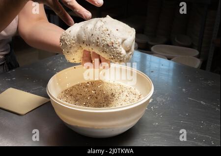 Nahaufnahme der weiblichen Hände einer Bäckerin, die den Teig in eine Schüssel mit Samen und Getreide eintaucht, bevor sie in einer Backstube hausgemachtes Brot backen Stockfoto