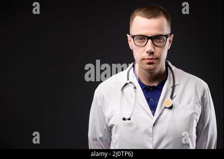 Ein Studioporträt eines attraktiven jungen kaukasischen Arztes in weißem Mantel und Brille mit Stethoskop um den Hals. Ständer Stockfoto