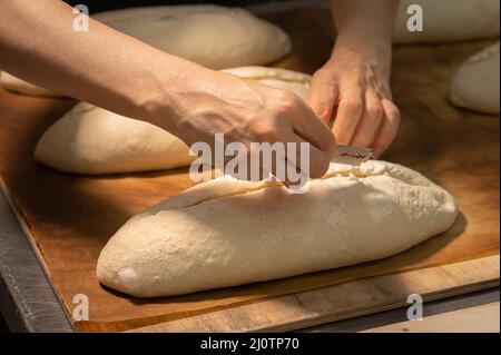 Nahaufnahme der weiblichen Hände einer Bäckerin, die einen Laib Teig mit einer Klinge in einen Laib Brot schneidet, bevor sie im Ofen backen. Craft cr Stockfoto