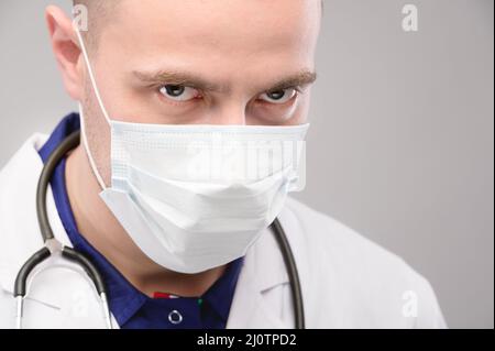 Der junge kaukasische, wütende Arzt in Maske und Robe blickt unheimlich in die Kamera. Impfverschwörung Stockfoto