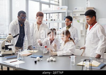 Eine große Gruppe verschiedener Kinder trägt Laborkittel im Chemieunterricht, während sie wissenschaftliche Experimente genießen Stockfoto