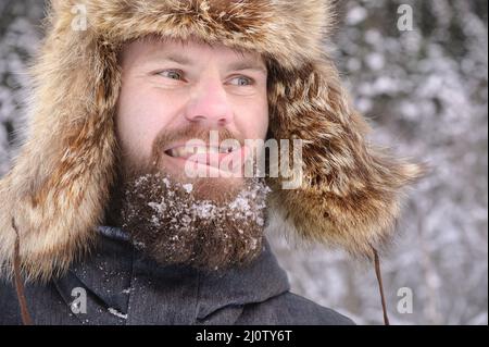 Porträt eines bärtigen Mannes in einem pelzgroßen Hut vor dem Hintergrund eines schneebedeckten Waldes. Lächelnder Mann im Wald. Anzeigen Stockfoto