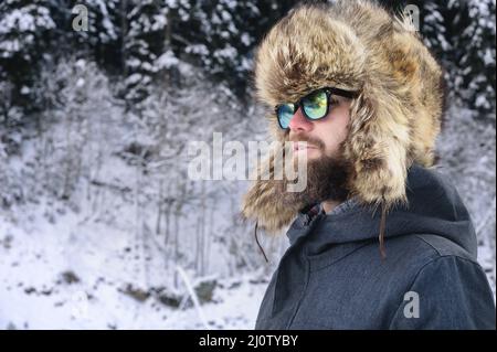 Porträt eines attraktiven jungen bärtigen Mannes in einem großen Pelzhut und Sonnenbrille gegen den schneebedeckten Wald in den Bergen Stockfoto
