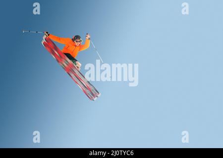 Stylische Skifahrer in orangefarbener Jackenkappe und Sonnenbrille in Jump machen den Trick isoliert auf einem blauen Hintergrund Stockfoto