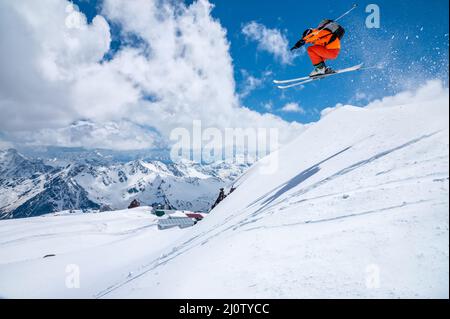 Ein männlicher Skifahrer in einem orangefarbenen Anzug fliegt in der Luft, nachdem er an einem sonnigen Tag hoch in den kaukasischen Bergen von einem Schneepfich gesprungen ist Stockfoto